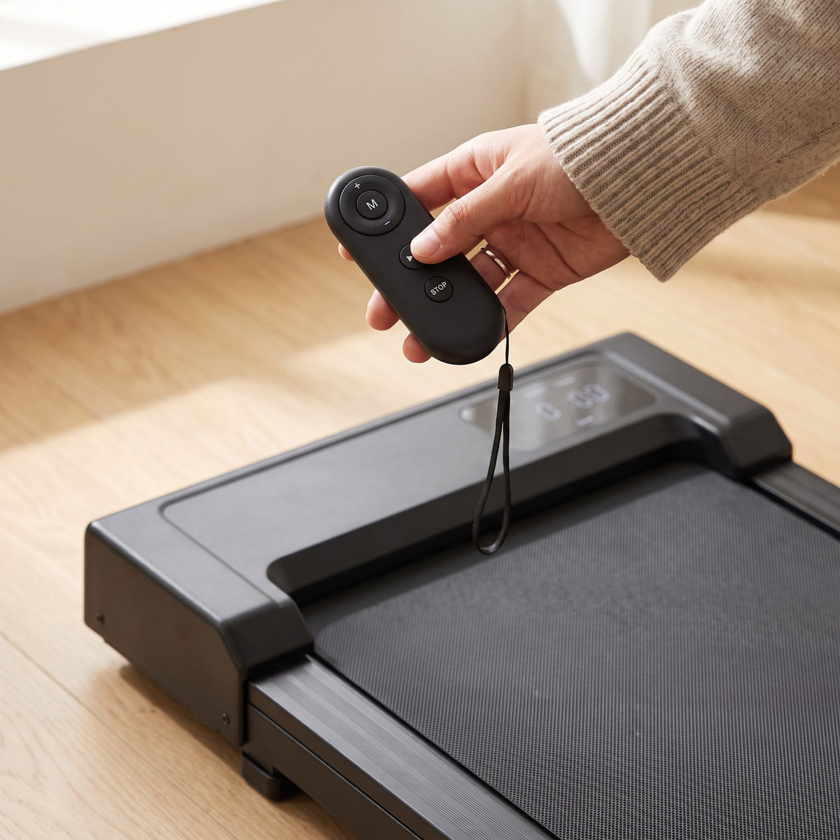 Hand holding the walking pad's remote control, with mode, start/pause and stop buttons, above the pad's display